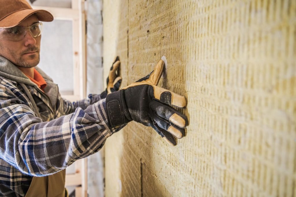 A worker in a plaid shirt and gloves applies insulation panels to a textured yellow wall in a construction site.