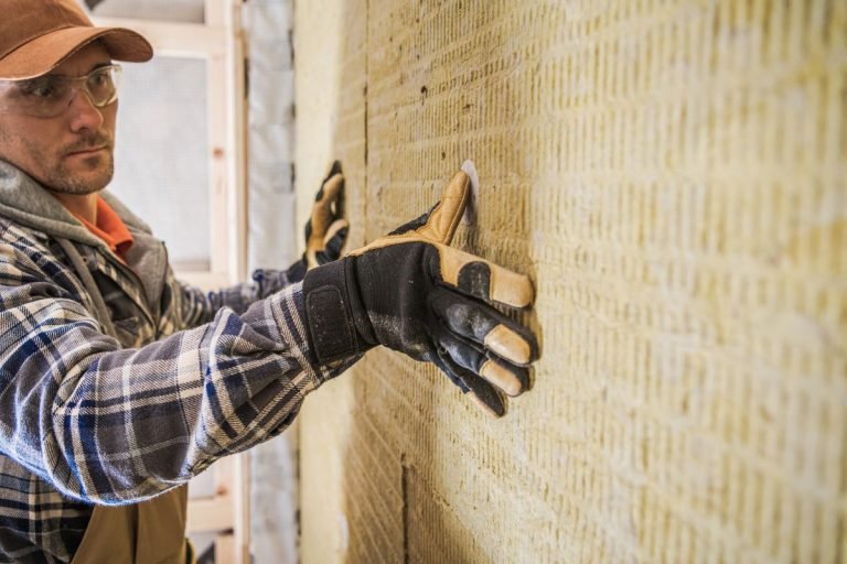 A worker in a plaid shirt and gloves applies insulation panels to a textured yellow wall in a construction site.