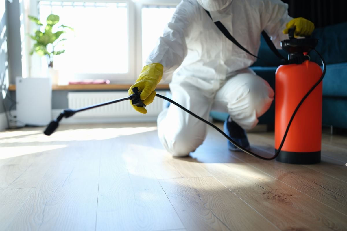 A person in protective gear sprays a floor with a pest control device, focused on ensuring a safe and thorough treatment indoors.